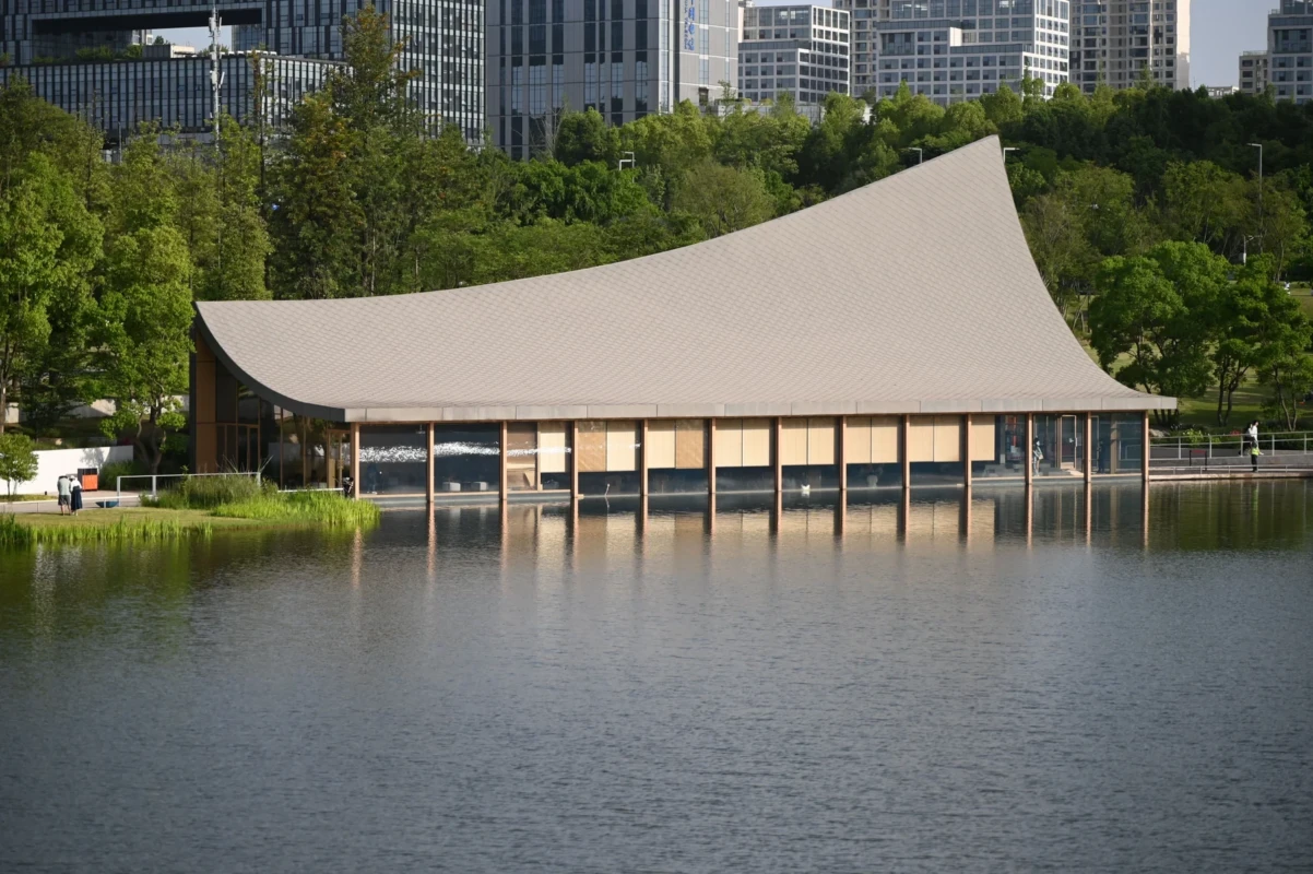 A contemporary curved-roof pavilion by an urban lake, framed by trees and a waterside approach.