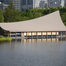 A contemporary curved-roof pavilion by an urban lake, framed by trees and a waterside approach.